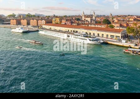 Ein kleines Flusskreuzfahrtschiff legt an einem Hafen in Venedig an, mit den großen Kreuzfahrtschiffen, die in der Ferne hinter dem Hauptschiffhafen zu sehen sind. Stockfoto