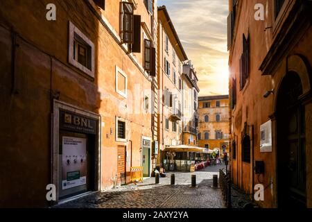 Eine schmale Gasse mit hohen Wänden auf beiden Seiten führt zu einem kleinen Platz mit einem Straßencafé, während die Sonne in Rom, Italien untergeht. Stockfoto