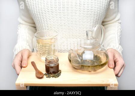 Ein Mädchen in einem weißen Pullover hält ein Holztablett mit einer Glaskanne, einem Becher, einem Löffel und einem Glas Honig. Oolong-Tee wird in der Teekanne gebrüht. Stockfoto