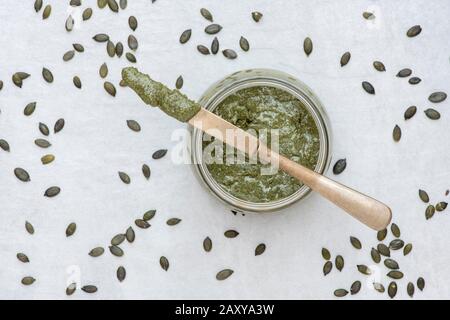 Hausgemachte vegane Kürbissaatbutter in einem kilner Glas mit einem Messer, das von einigen Kürbiskernen auf weißem Grund umgeben ist Stockfoto