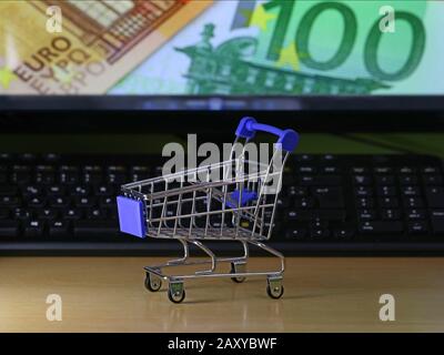 Einkaufswagen auf Holzschreibtisch mit Euro-Banknoten-Hintergrund auf dem Bildschirm, Konzept des Online-Einkaufs Stockfoto