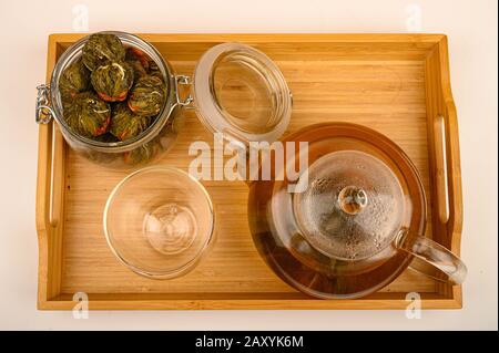 Blumentee, der in einer Glaskanne, einem Glas und einem Glas mit Blumenkugeln auf einem Holztablett auf weißem Hintergrund gebrüht wird. Nahaufnahme Stockfoto