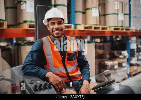 Der gutaussehende Ingenieur trägt eine Schutzuniform und einen Hut, der freudig zur Kamera lächelt, die im Gabelstapler sitzt, während er an der Lagerung arbeitet Stockfoto