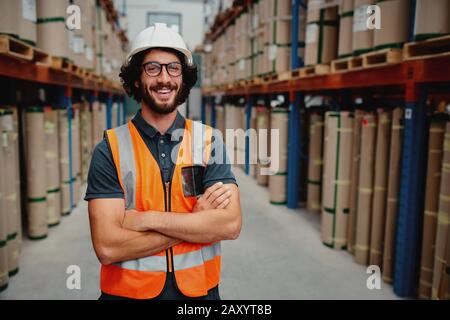 Fröhlicher Unternehmer für männliche Arbeiter in Uniform mit weißem Hardhat und mit Armen überzogen stehend aus der Lagerregalanordnung mit Brille Stockfoto