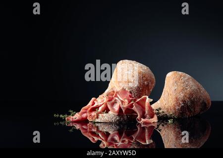 Prosciutto mit Ciabatta und Thymian auf schwarzem reflektierendem Hintergrund. Kopierbereich. Stockfoto