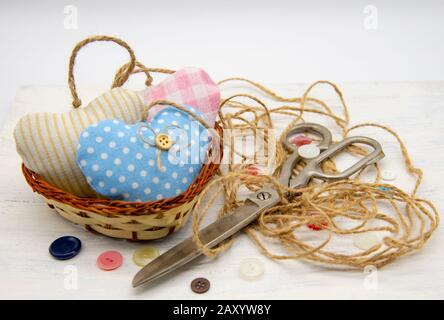 Ein Korb voller Herzen, genäht aus Stofffetzen, neben großen Scheren des Retro-Schneiders, grobem Faden und bunten Knöpfen. Stockfoto