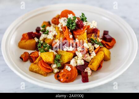 Weiße Keramikplatte mit geröstetem Gemüse (Kartoffel, Kürbis, Karotte, Rote Bete), frischer Petersilie und Fetakäse. Stockfoto