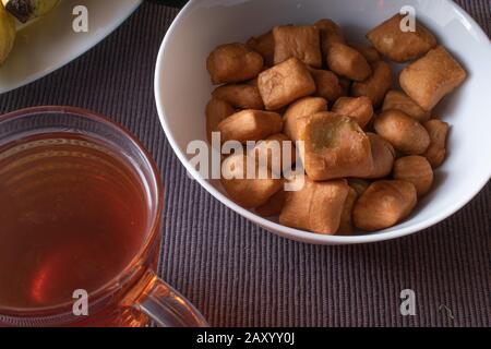 Nahaufnahme eines kleinen Mandazi in einer weißen Schüssel mit einer Tasse schwarzem Tee. In Kenia ist dies ein sehr beliebter Snack. Stockfoto