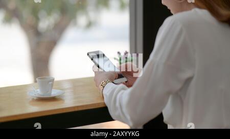 Zugeschnittene Aufnahme einer Frau, die auf dem Smartphone getextet wird, während sie in einem einfachen Café neben dem Fenster sitzt Stockfoto