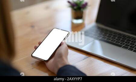 Zugeschnittene Aufnahme einer Frau, die ein leeres Bildschirm-Smartphone mit Laptop auf Holzschreibtisch hält Stockfoto