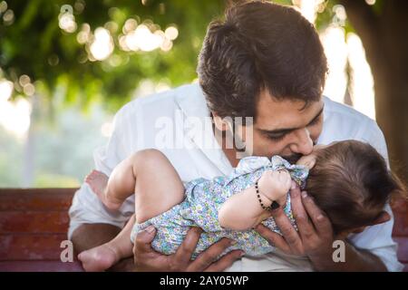 Porträt eines glücklichen indischen Vaters, der ihre Tochter hält und küsst, während er auf der Parkbank sitzt. Stockfoto