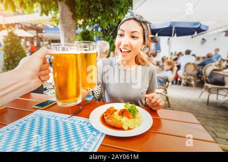 Fröhliches Asien-Mädchen clinks ein Becher bayerischen Bieres in Biergarten. Traditioneller deutscher Imbiss mit Wurst- und Kartoffelsalat Stockfoto