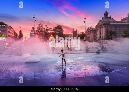 August 2019, München, Deutschland: Kind hat Spaß und läuft bei einem warmen Abend in München um einen Brunnen. Bewegungsunschärfe Stockfoto