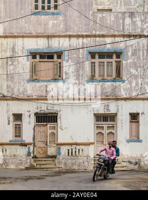 DIU, Indien - Dezember 2018: Zwei Männer radeln eine Straße an einem alten verwitterten Wohnhaus mit malerischen Fenstern und Holztüren vorbei. Stockfoto