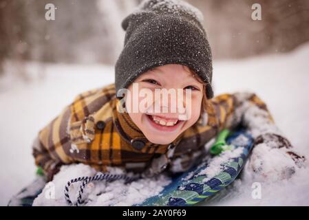 Porträt eines lächelnden Jungen, der auf einem Schlitten im Schnee liegt, Wisconsin, USA Stockfoto