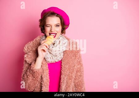 Foto der hübschen Dame Reisenden halten frische Croissant flirty Blink Eye Morgen-Snack im Ausland Beißteig tragen Freizeit stylische Fellmantel Schal Beret Kappe Stockfoto