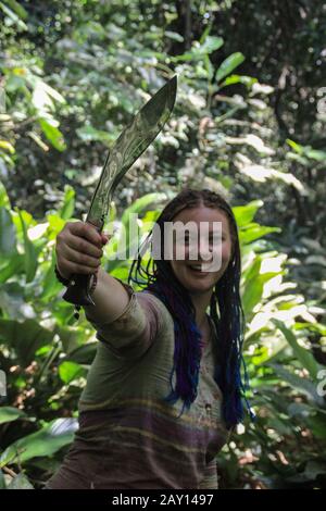 Ein plump junges weißes Mädchen mit blauen Ferkelhaaren im Dschungel, das eine Machete hält. Stockfoto