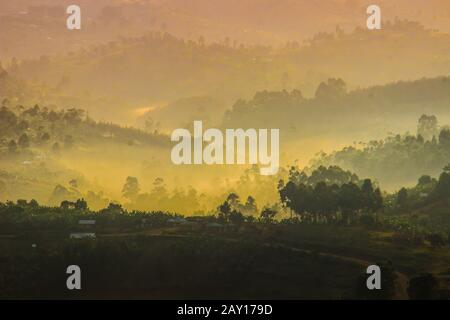 Sanftes gelbes Licht und leichter Nebel über den Hügeln auf der Landseite mit traditionellen Häusern und der tropischen Natur von Uganda an der Grenze zur Con Stockfoto