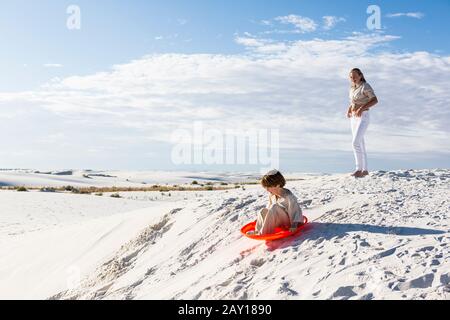 Kinder, die in Sanddünen spielen, eine auf einem orangefarbenen Schlitten. Stockfoto