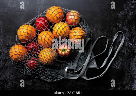 Hochwinkelige Nahaufnahme von roten Äpfeln und Orangen in grauer Netztasche auf schwarzem Grund. Stockfoto
