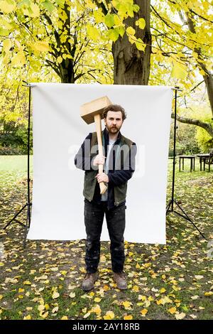 Porträt eines bärtigen Mannes, der Holzklotz vor weißem Hintergrund in einem Garten hält und Kamera betrachtet. Stockfoto