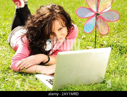 Fröhliches junges Mädchen, das in der Natur auf grünem Gras liegt und an einem Laptop arbeitet Stockfoto