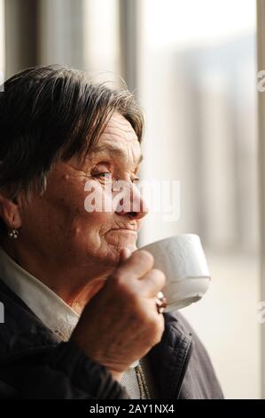 Porträt der älteren Frau, die durch Fenster schaut und Kaffee trinkt Stockfoto