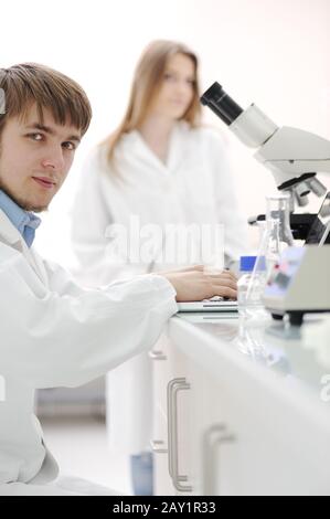 Junger männlicher Forscher mit Mikroskop und Schreiben von Notizen auf Laptop im Labor Stockfoto