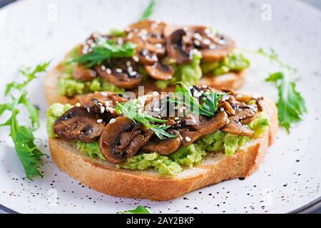 Bruschetta. Sandwich mit Avocado, gebratenen Pilzen, Sesam auf einem weißen Tisch. Vegetarisches Essen. Veganes Menü. Stockfoto