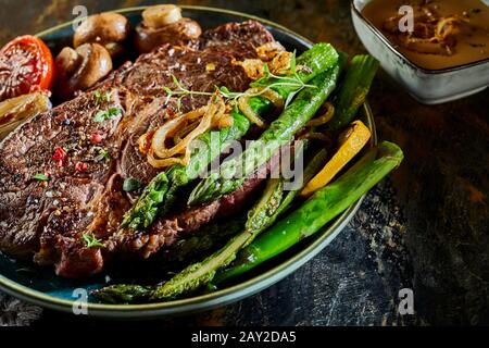 Frische grüne Spargelspeere mit einem dicken saftigen gegrillten Rindersteak, Champignons und Tomaten für ein saisonales Gourmet-Abendessen Stockfoto