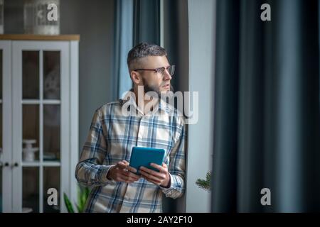 Bärtiger junger Mann mit einer Tablette, die am Fenster steht. Stockfoto