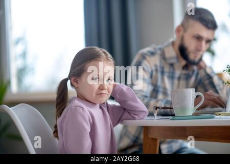 Kleines Mädchen am Tisch sitzend traurig lächelnd, Papa beschäftigt. Stockfoto