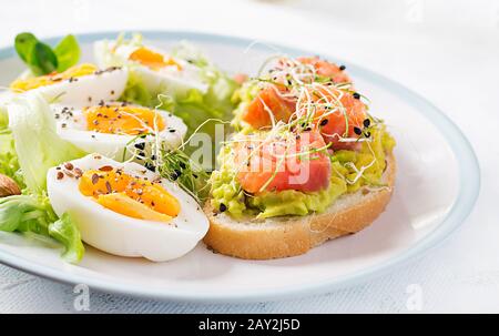 Frühstück. Gesundes, offenes Sandwich auf Toast mit Avocado und Lachs, gekochten Eiern, Kräutern, chia-Samen auf weißem Teller mit Kopierraum. Gesundes Protein fo Stockfoto