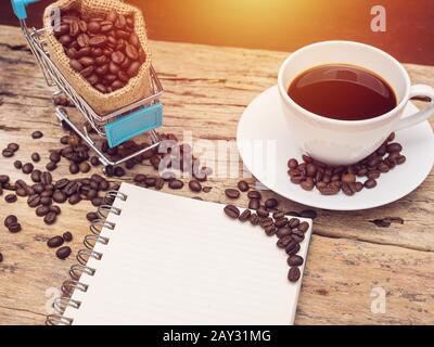 Leeres Buch mit Platz zum Kopieren und einer Tasse frischen schwarzen heißen Kaffees mit Einkaufswagen voller Kaffeebohne über Holzgrund. Kaffeerezept, Barista Stockfoto