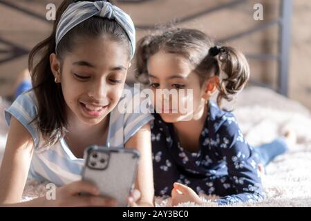 Zwei dunkelhäutige junge Mädchen, Schwestern, liegen im Bett und schauen sich ein Smartphone vor dem Hintergrund der Sonne vom Fenster. Persische Mädchen Stockfoto