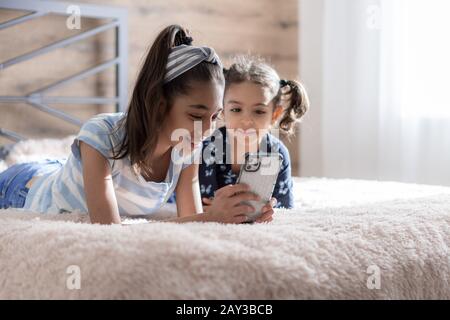 Zwei dunkelhäutige junge Mädchen, Schwestern, liegen im Bett und schauen sich ein Smartphone vor dem Hintergrund der Sonne vom Fenster. Persische Mädchen Stockfoto