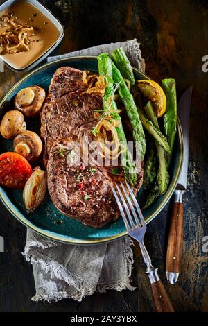 Portion gegrilltes Rindersteak mit frischen grünen Spargelspeeren, gerösteten Zwiebeln, Champignons und Tomaten, garniert mit Kräutern, die oben unten am Tisch betrachtet werden Stockfoto