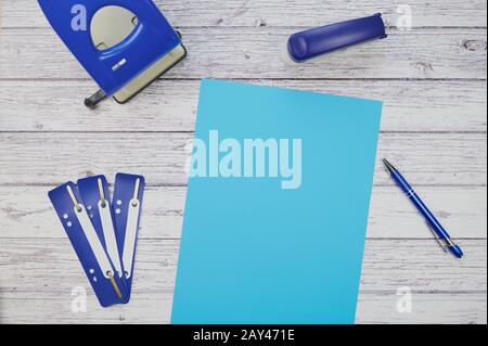 Blaues Blatt Papier auf weißem Holzhintergrund, Stanzer, Hefter, Stift und Aktenstreifen liegen um ihn herum Stockfoto