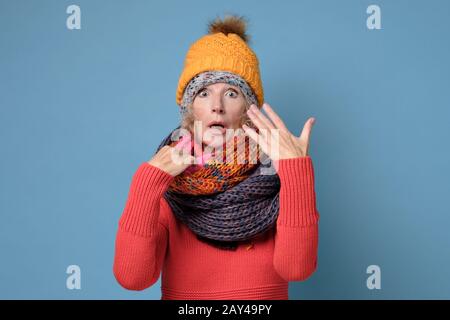 Seniorin in Hüten und Schals ist mit Schweiß bedeckt, weil sie zu viel Kleidung trägt, die an der blauen Wand isoliert ist. Kaltwetterkonzept Stockfoto