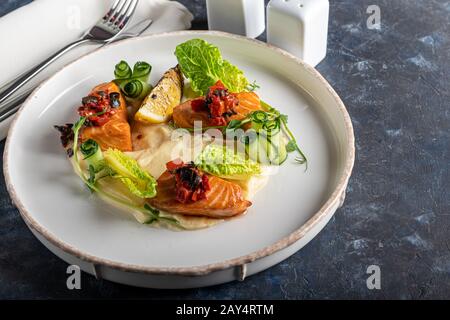 Sellerie und Kartoffelpüree mit roten Fischstücken, garniert mit Kräutern und frischen Gurken. Auf einer weißen Platte mit Zitronenscheiben. Auf einem Holztisch. Kopieren Stockfoto