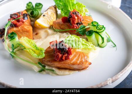 Sellerie und Kartoffelpüree mit roten Fischstücken, garniert mit Kräutern und frischen Gurken. Auf einer weißen Platte mit Zitronenscheiben. Auf einem Holztisch. Kopieren Stockfoto