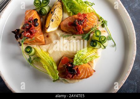 Sellerie und Kartoffelpüree mit roten Fischstücken, garniert mit Kräutern und frischen Gurken. Auf einer weißen Platte mit Zitronenscheiben. Auf einem Holztisch. Kopieren Stockfoto