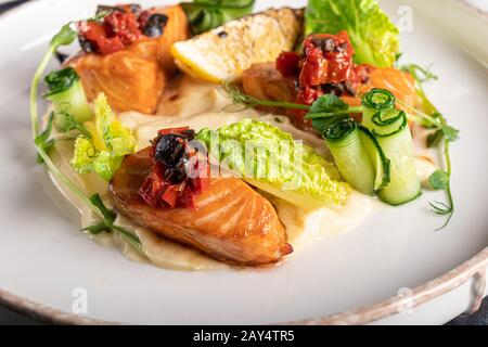 Sellerie und Kartoffelpüree mit roten Fischstücken, garniert mit Kräutern und frischen Gurken. Auf einer weißen Platte mit Zitronenscheiben. Auf einem Holztisch. Kopieren Stockfoto