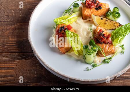 Sellerie und Kartoffelpüree mit roten Fischstücken, garniert mit Kräutern und frischen Gurken. Auf einer weißen Platte mit Zitronenscheiben. Auf einem Holztisch. Kopieren Stockfoto