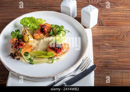 Sellerie und Kartoffelpüree mit roten Fischstücken, garniert mit Kräutern und frischen Gurken. Auf einer weißen Platte mit Zitronenscheiben. Auf einem Holztisch. Kopieren Stockfoto