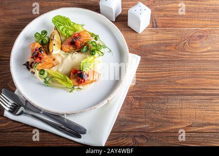 Sellerie und Kartoffelpüree mit roten Fischstücken, garniert mit Kräutern und frischen Gurken. Auf einer weißen Platte mit Zitronenscheiben. Auf einem Holztisch. Kopieren Stockfoto