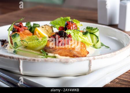 Sellerie und Kartoffelpüree mit roten Fischstücken, garniert mit Kräutern und frischen Gurken. Auf einer weißen Platte mit Zitronenscheiben. Auf einem Holztisch. Kopieren Stockfoto