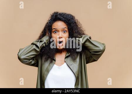 Die angetromte afroamerikanische Frau hält beide Hände am Kopf, öffnet weit den Mund, ist mit weißem T-Shirt und Lederhemd bekleidet und überrascht von etwas, Stockfoto