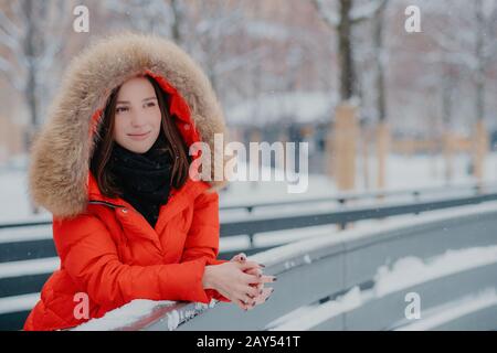 Außenaufnahme der nachdenklichen europäischen Frau trägt rote Winterjacke mit Kapuze am Kopf, lehnt sich daher an, blickt penibel in die Ferne, genießt guten Frost Stockfoto