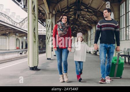 Fröhliche Brunette-Frau trägt roten Pullover und Schal, kleines Kind in übergroßem Pullover, gutaussehender Mann trägt Koffer, kehrt von Reise zusammen, Eilboten Stockfoto